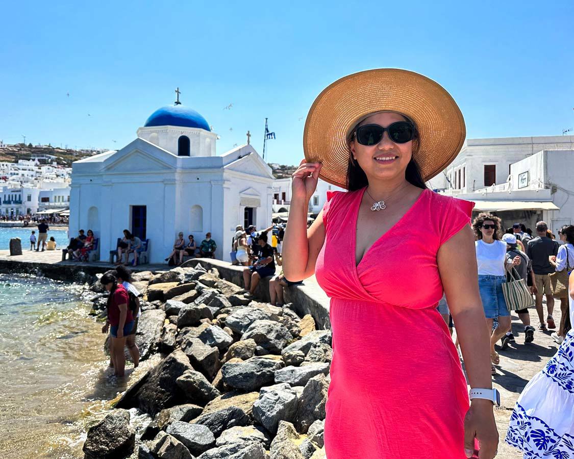 A woman in a pink dress smiles in front of the Agios Nikolaos Church in Old Chora, Greece