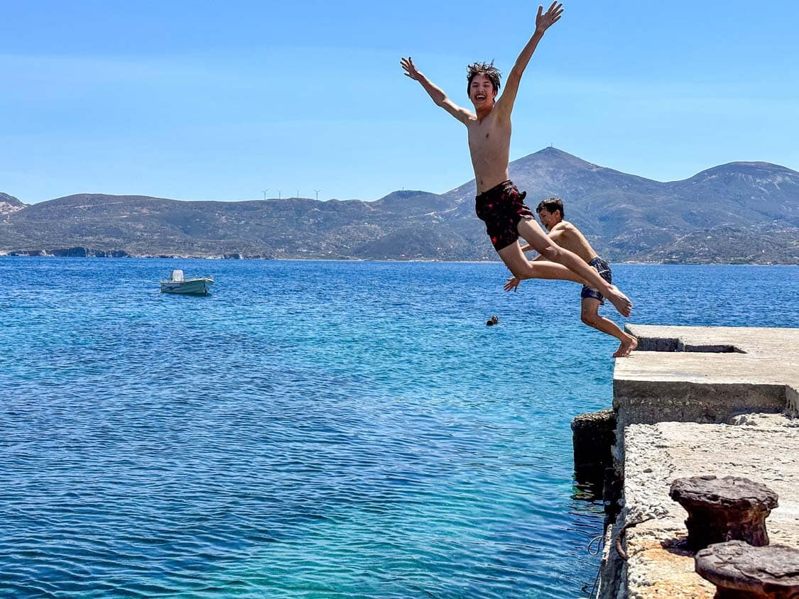 Two boys jump from a pier into the blue waters of Klima on Milos, Greece