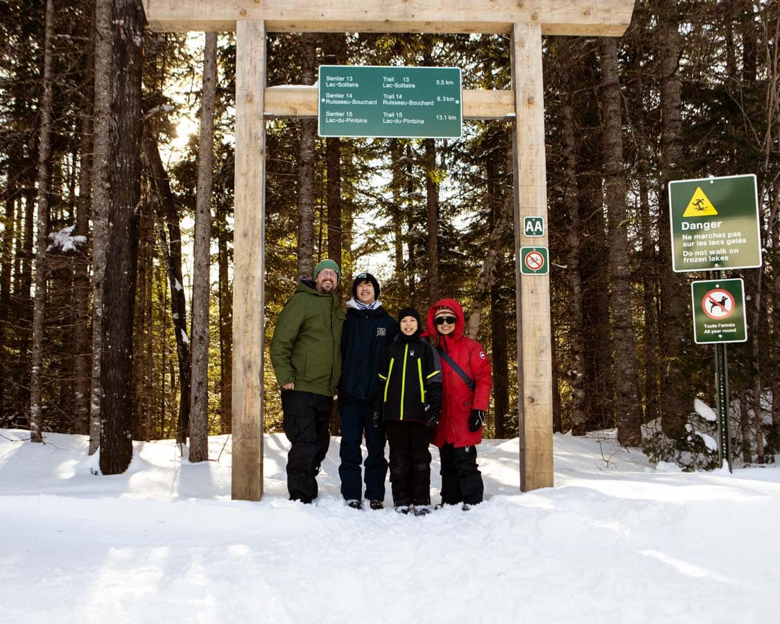 A family poses under the trail sign for the Lac Solitaire Trail during winter in La Mauricie National Park