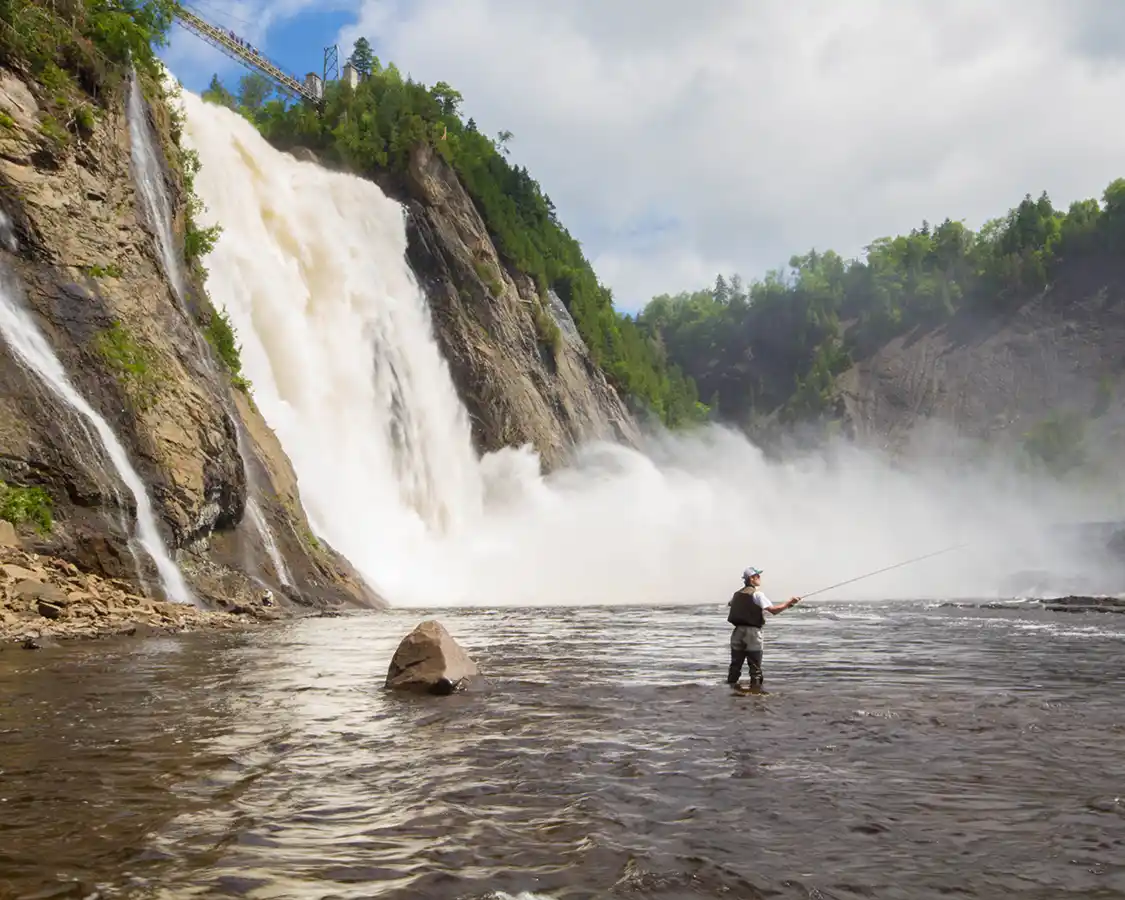 A man fly fishing at the base of a massive waterfall near Quebec City