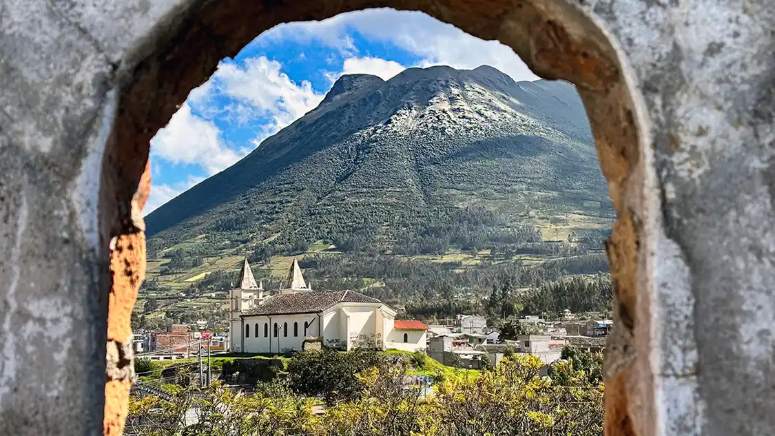 A small town and church lie next to a lush mountain as seen through a stone window frame in Otavalo, Ecuador.
