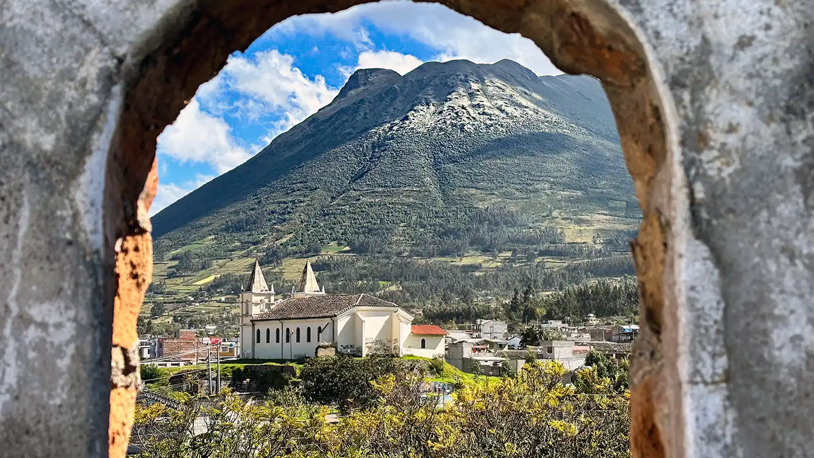 A small town and church lie next to a lush mountain as seen through a stone window frame in Otavalo, Ecuador.