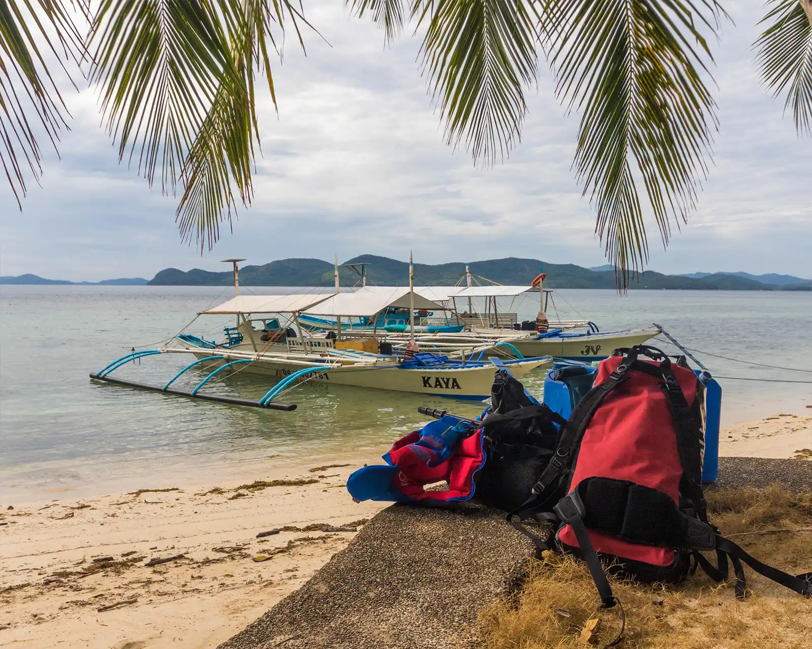 A collection of backpacks and waterproof bags sit under a palm tree on a beach in the Philippines