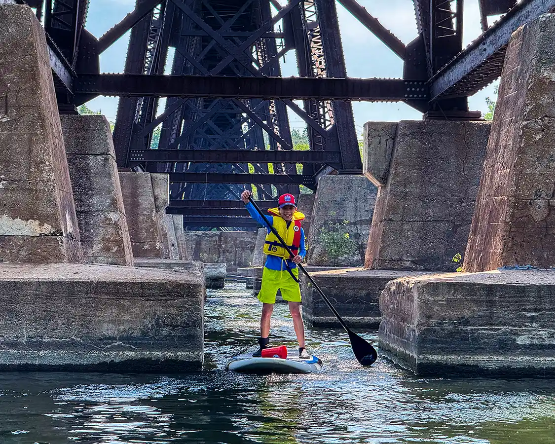 A boy navigates a stand up paddleboard through a train bridge on the Trent River in Trenton