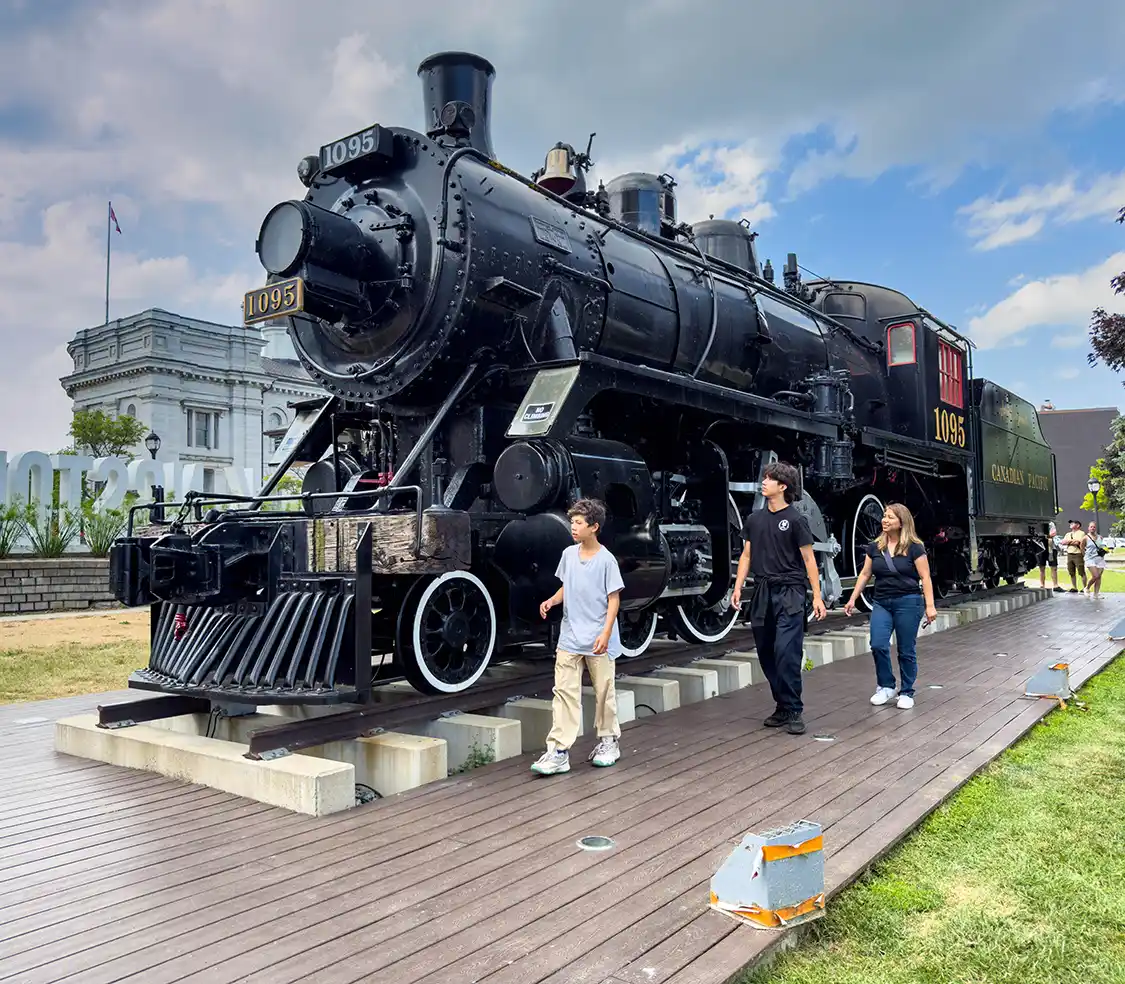 A mother and her two sons walk past a large train engine in Confederation Park, Kingston