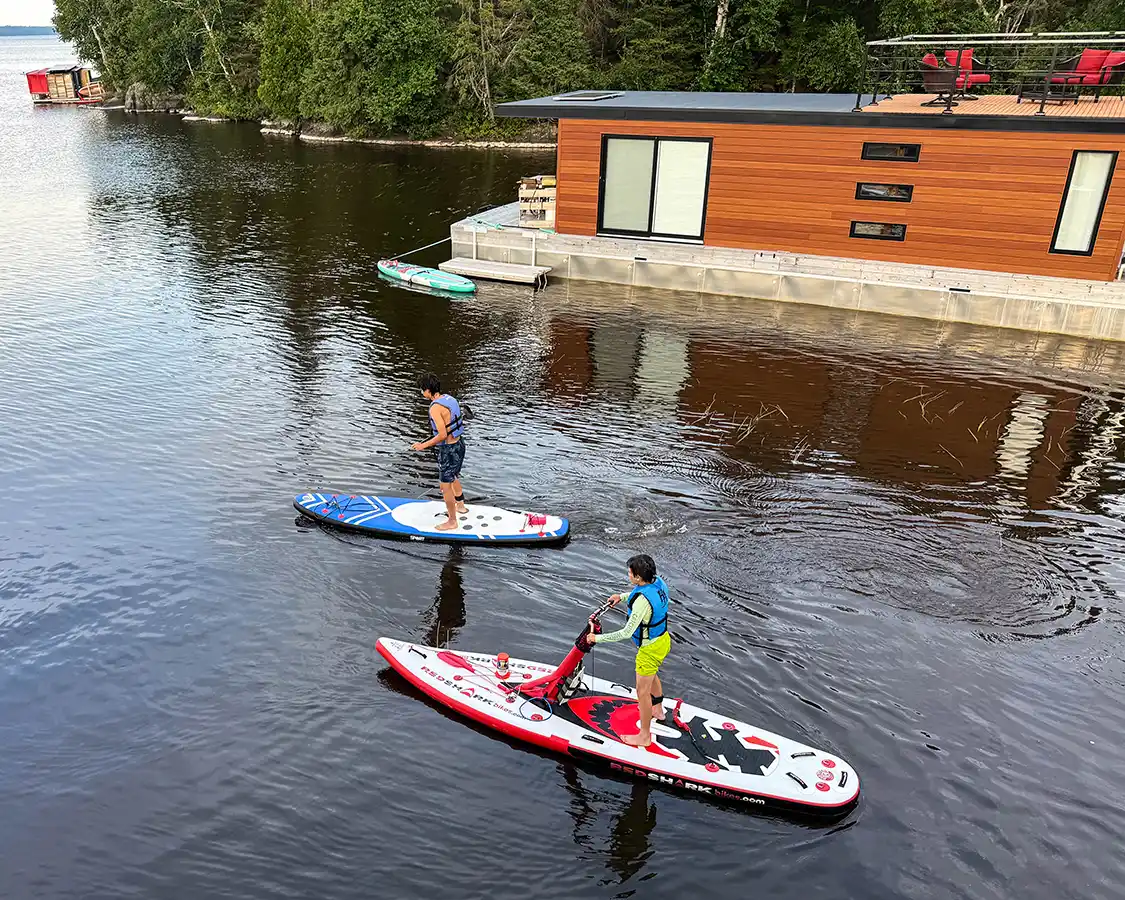 Two boys paddling stand up paddleboards by a floating cottage near Val d'Or Quebec