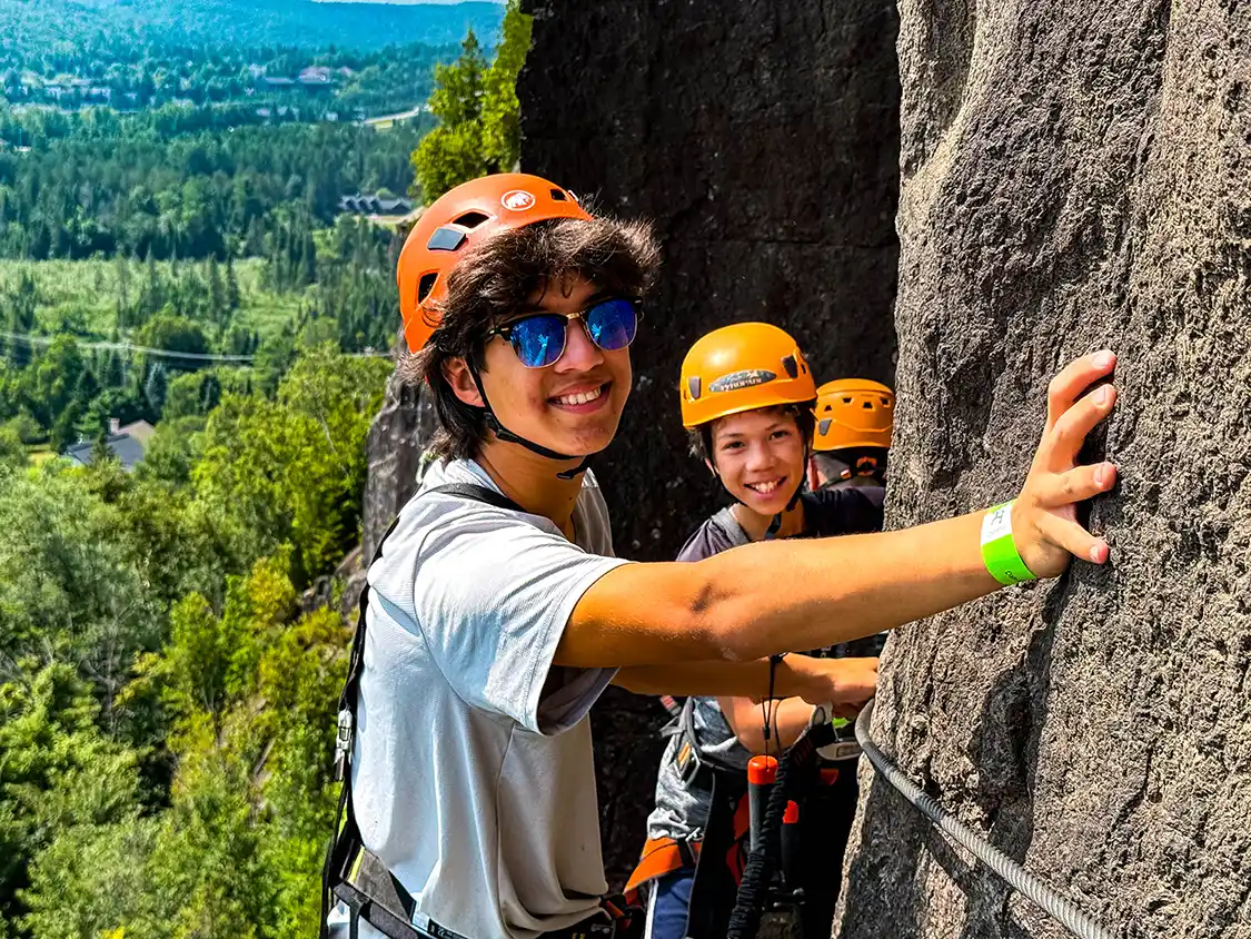 Two boys hang on the edge of a cliff on a Via Ferrata experience in Mont-Tremblant, Quebec during the summer