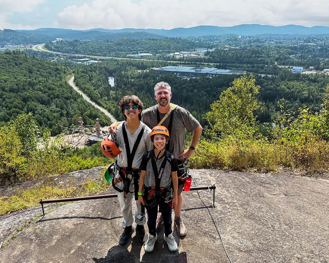 A father and his two sons smile at the top of a Via Ferrata overlooking the Laurentian Mountains in Quebec