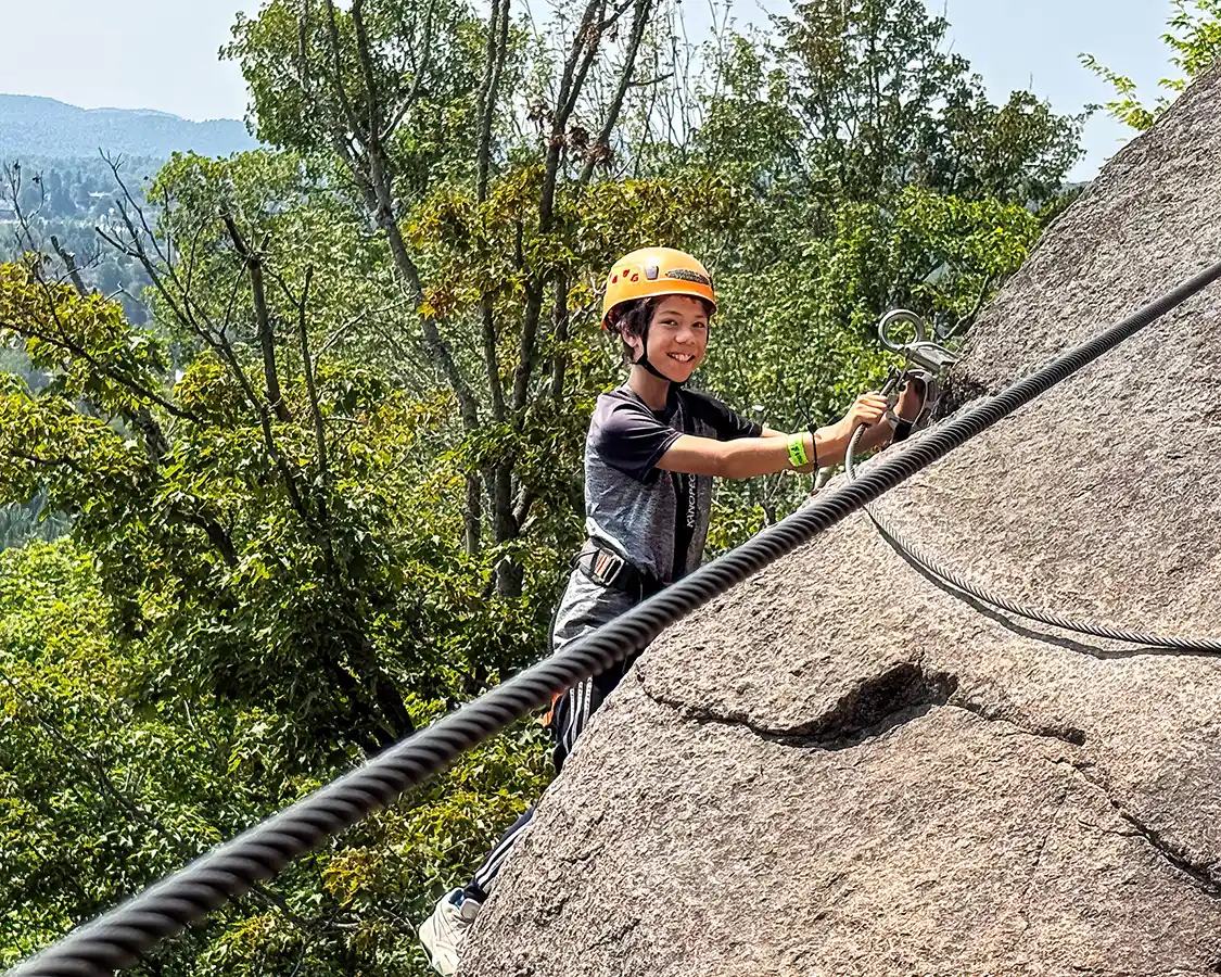 A boy smiles while hanging off the edge of a cliff on a Via Ferrata