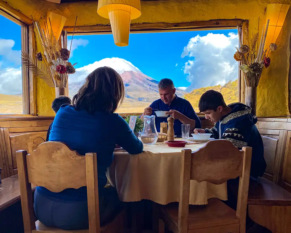 A family enjoys a dinner inside a rustic lodge in front of a window with views of Cotopaxi volcano