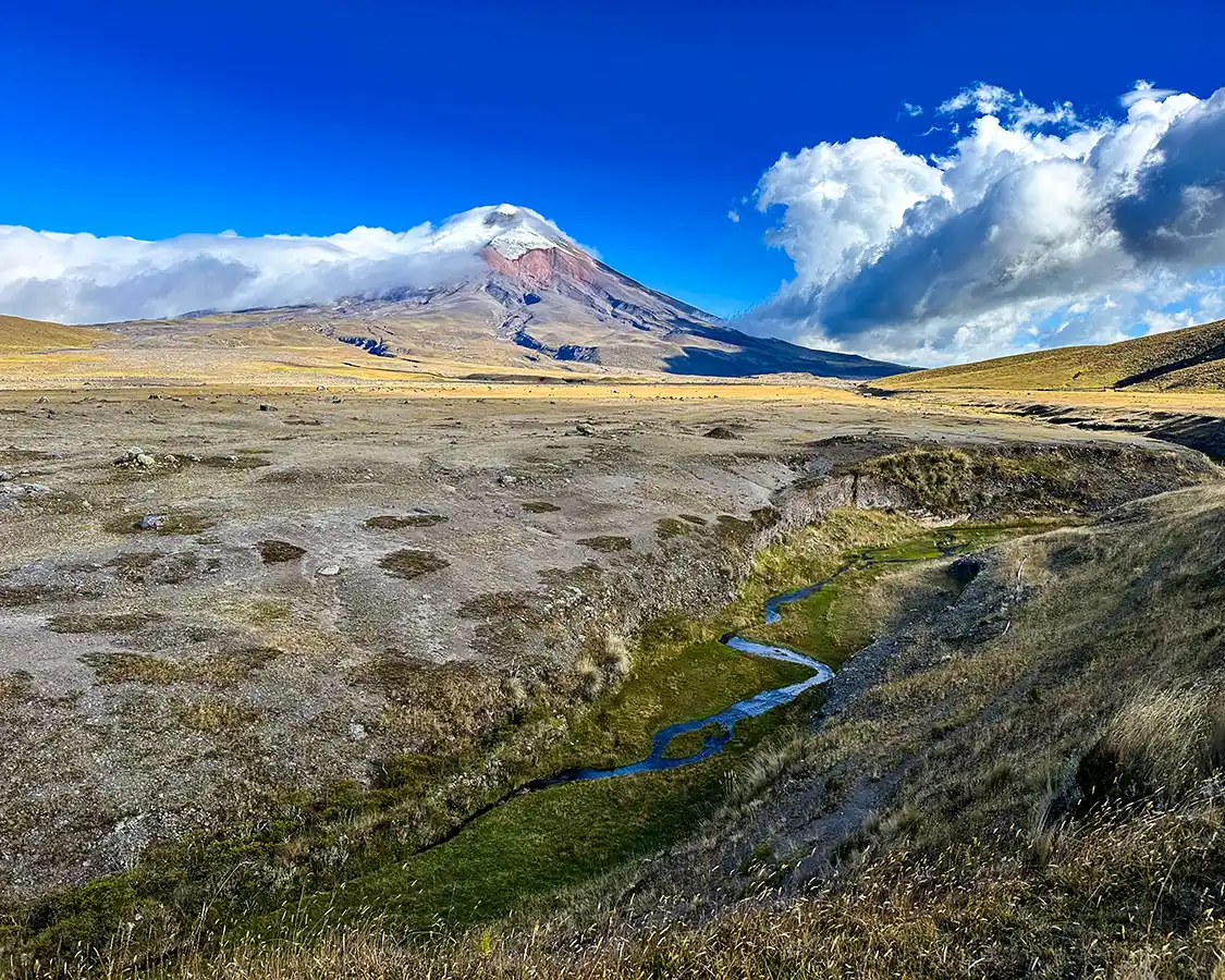A small stream winds through grasslands toward a cloud-tipped Cotopaxi volcano beneath deep blue skies