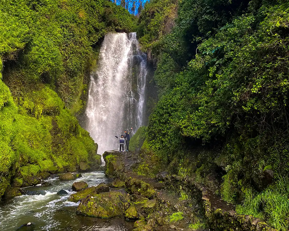 Two boys standing arm and arm and waving under a beautiful waterfall in Otavalo, Ecuador