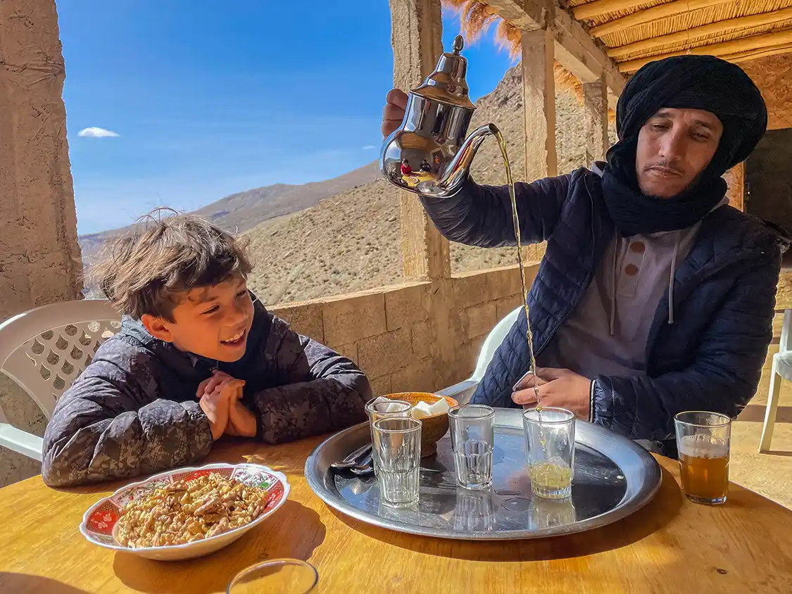 A Moroccan Berber man dramatically pouring tee while a young boy watches with a smile on his face