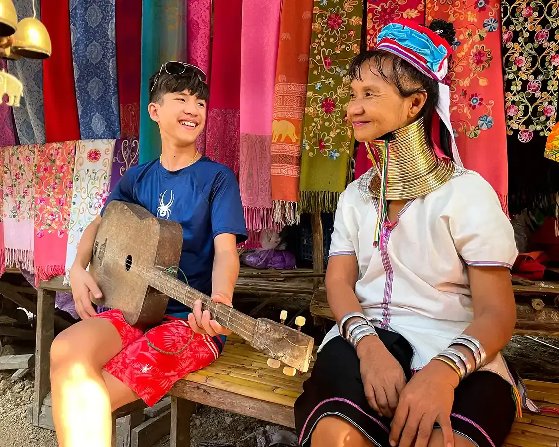 A boy holding a guitar smiles next to an elder from the Thailand Karen Hill Tribes