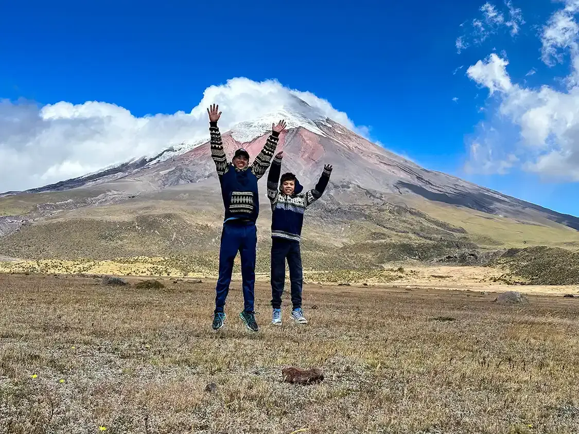Two boys in traditional sweaters jump in front of Cotopaxi Volcano