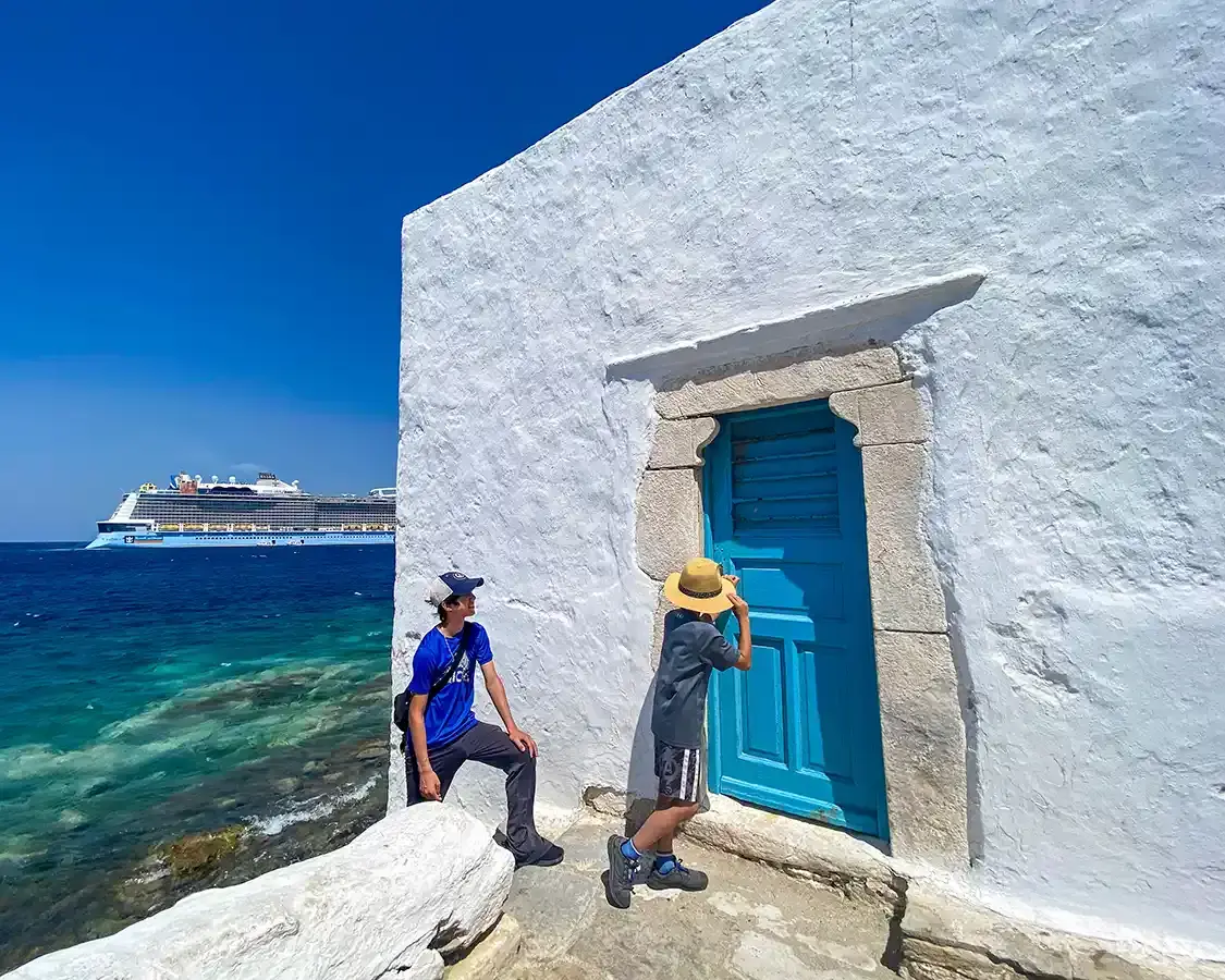Two boys walk toward a blue door with a cruise ship in the background in Mykonos Greece