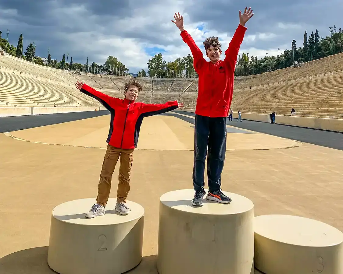 Two boys wearing red sweaters and leaf crowns stand atop the podium at the Olympic oval in Athens Greece